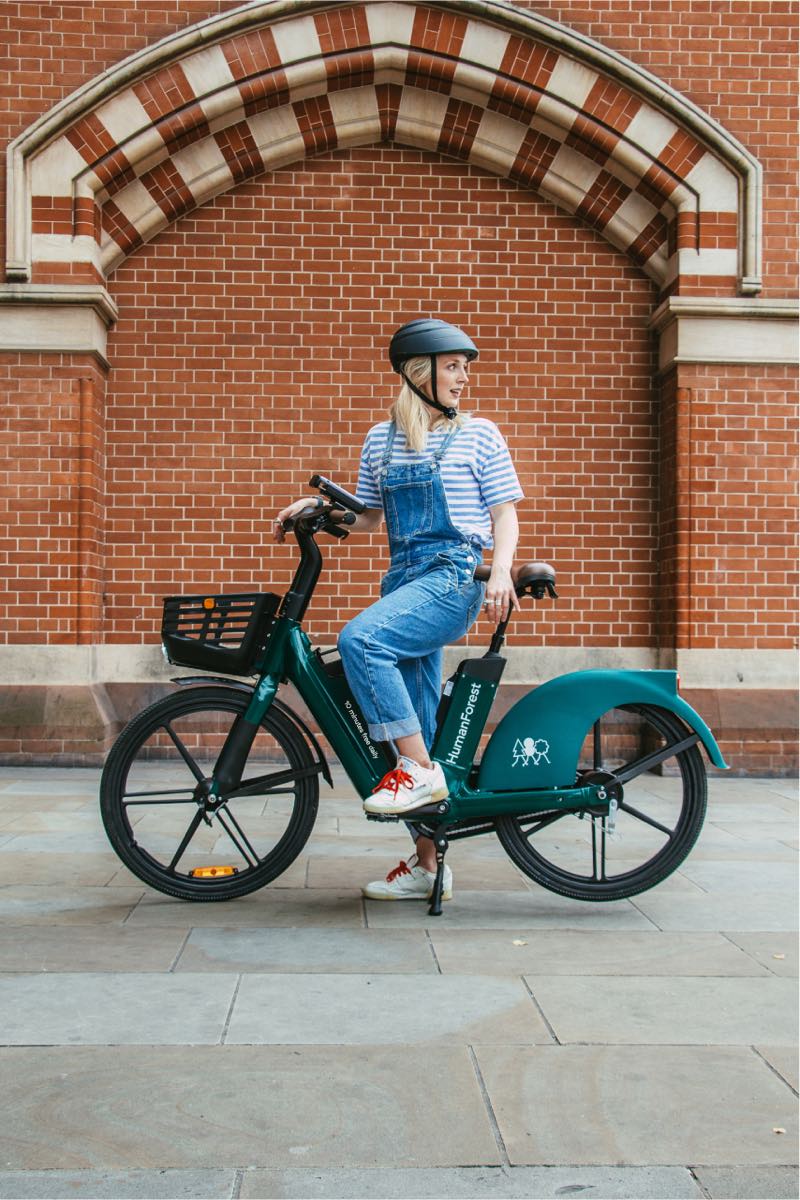A rider on a teal Human Forest shared e-bike in front of a red-brick London building.