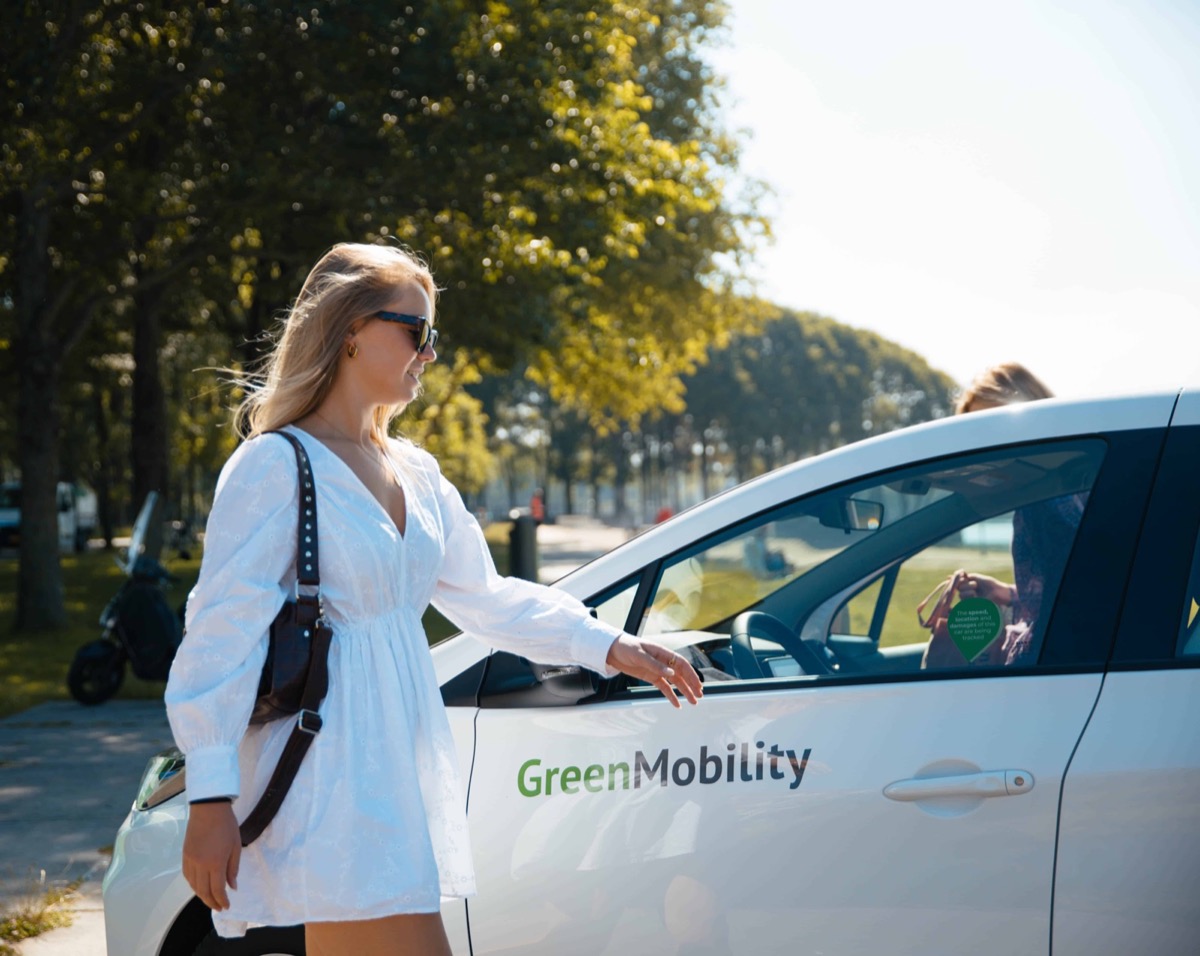 A rider walking up to a white GreenMobility free-floating car parked on a tree-lined city street.