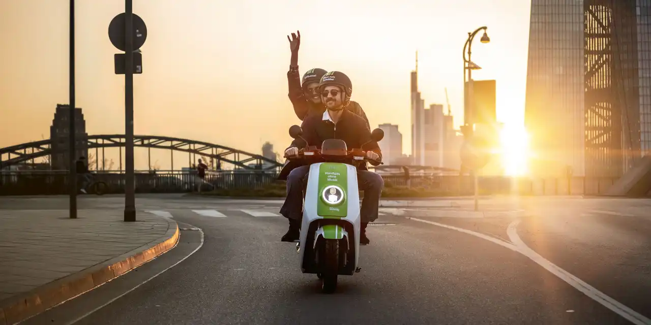 Two riders on a green Süwag2GO 'Frank-e' shared e-moped at sunset, with the Frankfurt skyline behind